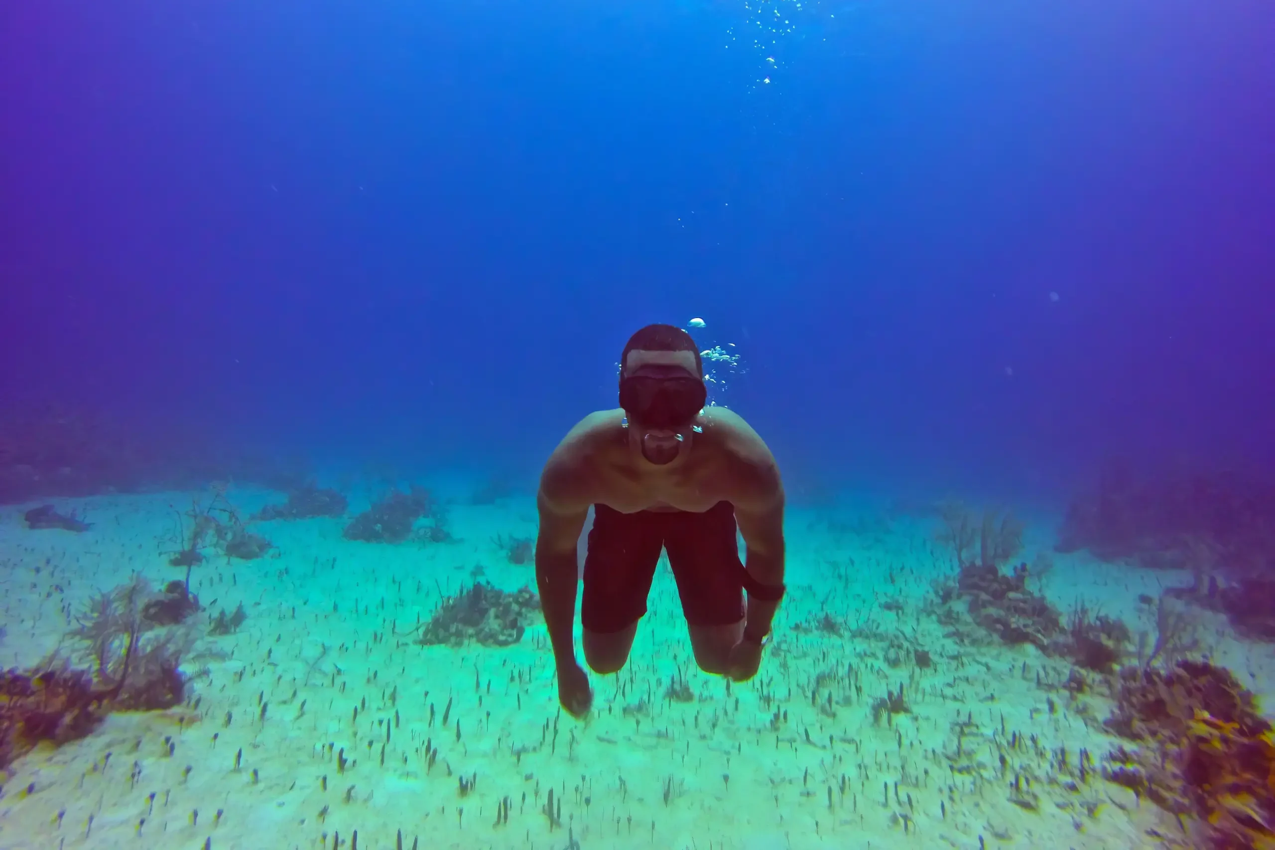 Person in a snorkel mask swims underwater above a sandy seabed, with bubbles rising into deep blue water.