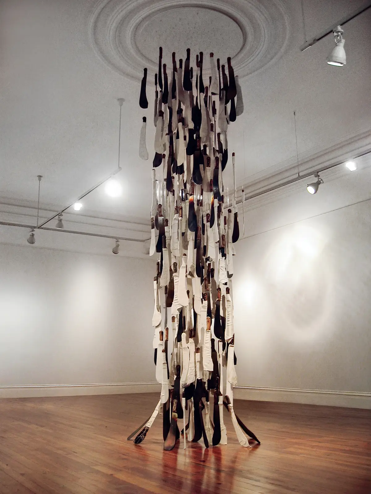 Gallery installation featuring a tall hanging cluster of paddle-shaped objects suspended from the ceiling in a white-walled room.