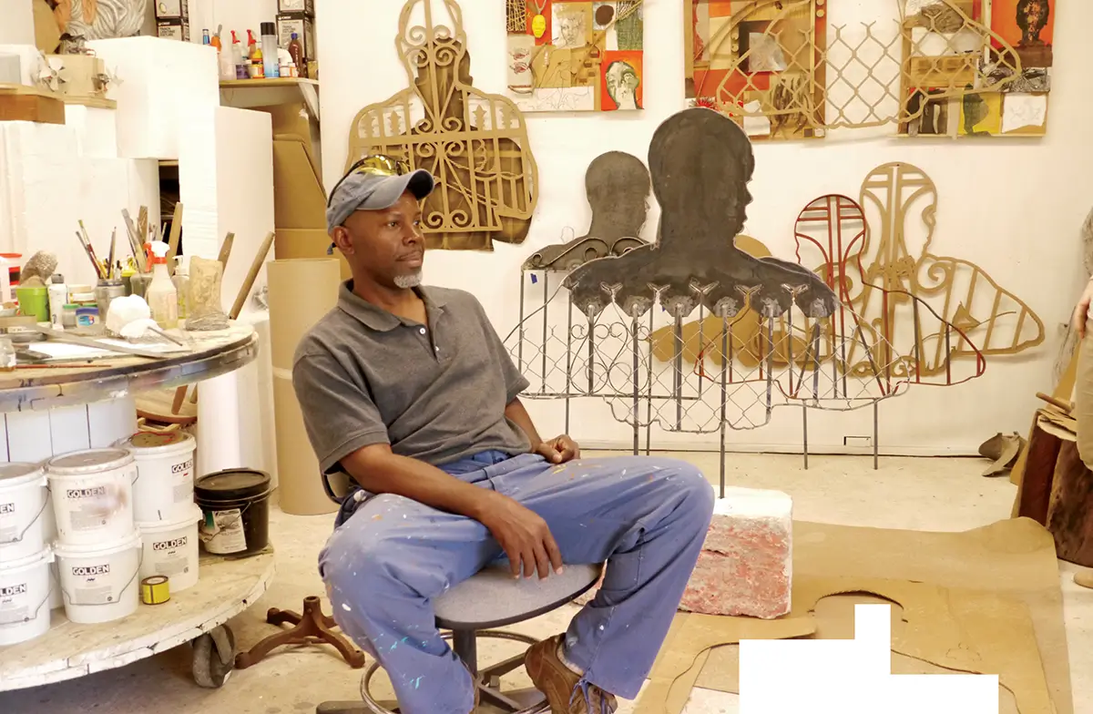 Artist seated in a studio surrounded by materials and large cutout silhouettes and metalwork forms leaning against the walls.