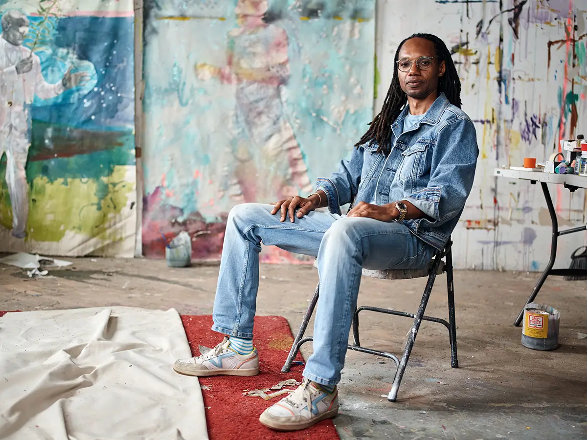 Artist Portrait in Studio Alt text: Person seated on a chair in a paint-splattered studio, wearing denim, with large canvases and abstract artwork in the background.